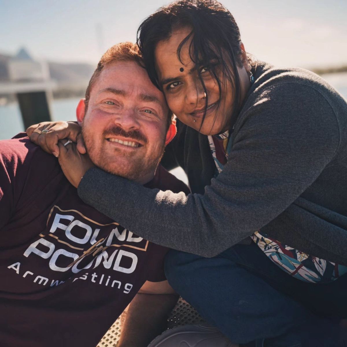 Chetna hugging Ryan by the water, Ryan wearing a Pound for Pound Armwrestling shirt